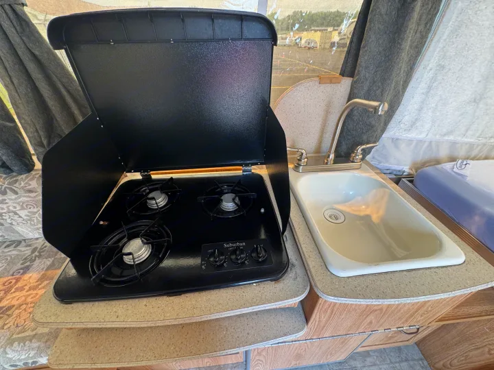 Compact RV rental kitchen with a black three-burner Suburban stove, wind guards open, beside a small sink and faucet.. Forest River Yearlying 4100 2008