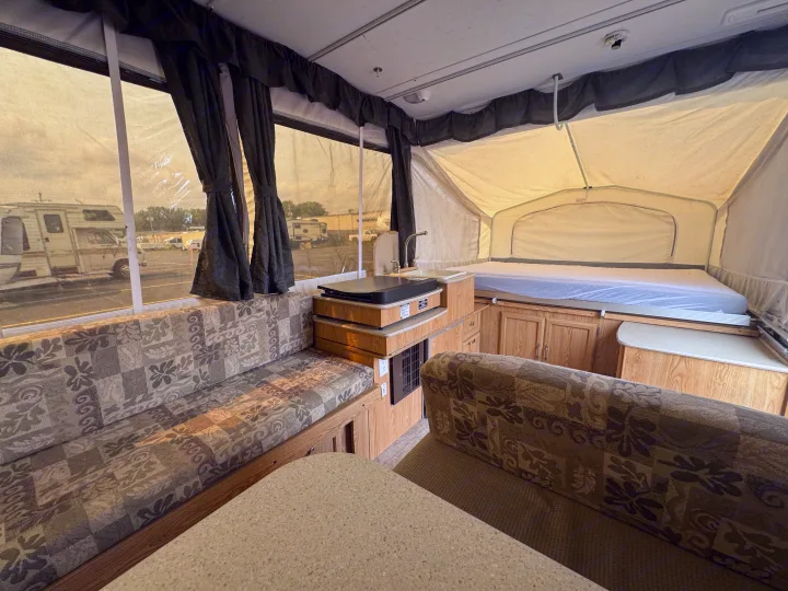 Interior of an rv rental pop-up camper: a patterned dinette, compact kitchen, and bed are visible. Outside, other RVs are parked.. Forest River Yearlying 4100 2008