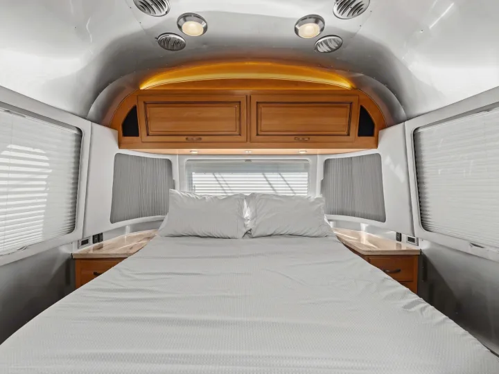 Cozy RV rental bedroom with a comfortable bed, overhead wooden cabinets, and windows with blinds in a metallic, modern interior.. Airstream Classic 2019