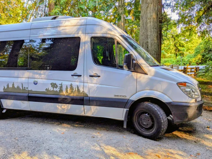 White camper van (RV rental) with tree decals, parked on gravel in a lush green forest, ready for adventures.. Mercedes Sprinter 2008