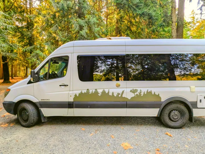 Stylish white Sprinter rv rental campervan, featuring a forest decal, parked amongst tall trees on gravel.. Mercedes Sprinter 2008