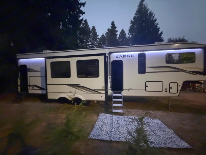 Spacious Sabre fifth-wheel RV rental at a nighttime campsite, featuring exterior LED lighting and an outdoor mat.. Forest River Sabre 2025