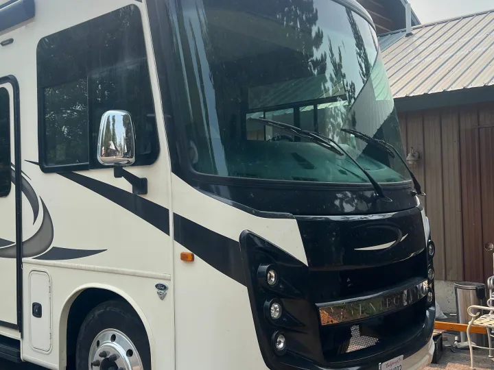 Close-up of a parked white and black motorhome, ideal for an RV rental. Shows the front, driver's side, windshield, and chrome mirror.. Entegra Coach Vision XL 2021