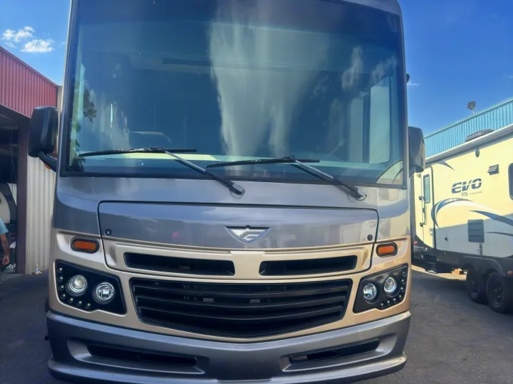 Detailed front of a grey and beige RV rental motorhome. Its large windshield reflects the sky, parked near a white travel trailer.. Fleetwood 35 K 2017