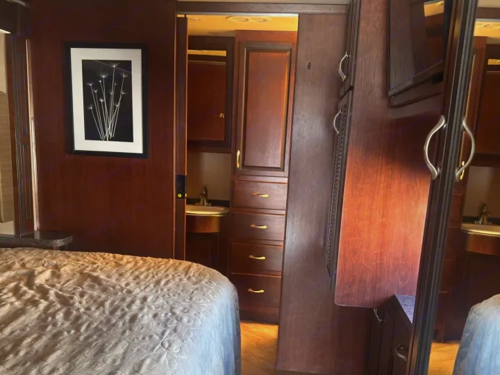 RV rental bedroom with a quilted bed, dark wood walls, and a glimpse of the bathroom sink through the open door.. Fleetwood 35 K 2017