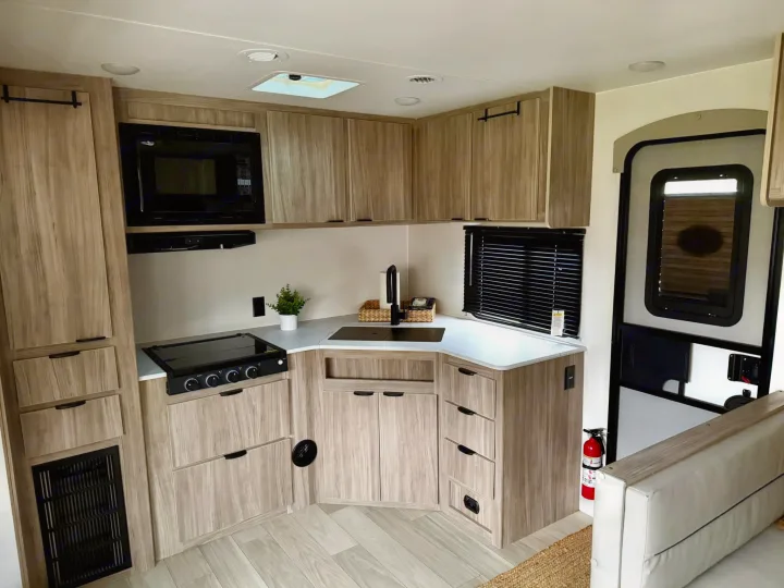 Functional RV rental kitchen. Light wood cabinets, black microwave, stovetop, and sink. Includes essential safety features.. Winnebago Access 2024