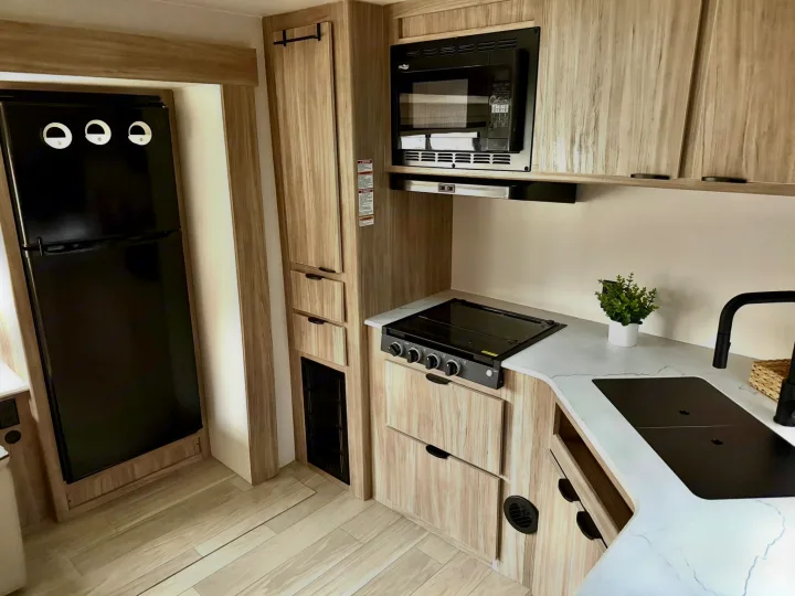 Bright RV rental kitchen with black fridge, microwave, stovetop, light wood cabinets, and modern flooring.. Winnebago Access 2024