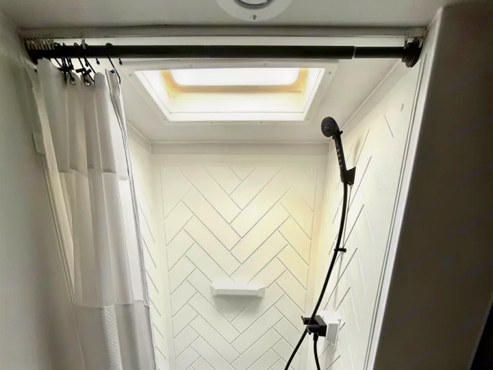 Bright and clean RV rental shower interior with white herringbone walls, skylight, and black shower fixtures for comfortable travel.. Winnebago Access 2024