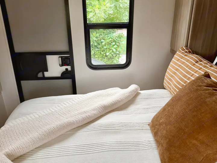 Cozy RV rental bedroom featuring a striped pillow, white textured bedding, and a window with a green nature view.. Winnebago Access 2024