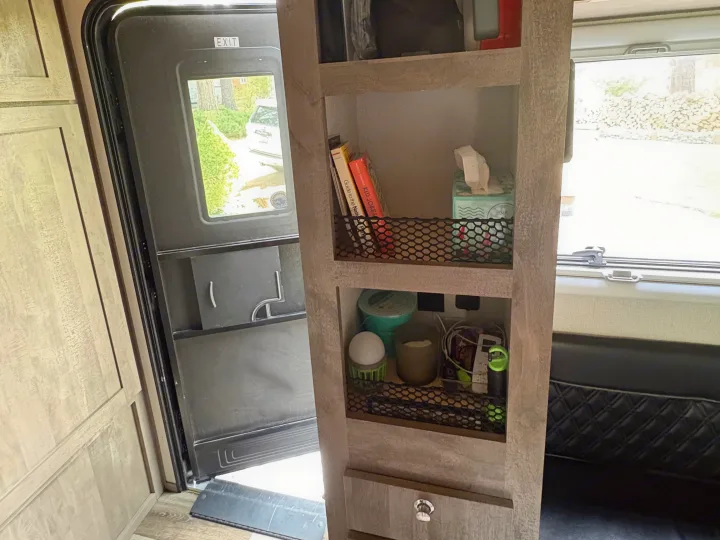 Inside an RV rental, an open door views outside, beside narrow shelves with books, tissues, and essentials for travel.. Imperial Outdoors XploreRV 2022