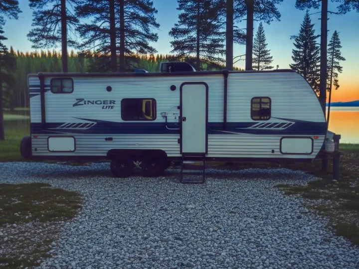 A Zinger Lite rv rental parked on gravel at a picturesque lakeside campsite with tall pine trees under a sunset sky.. Crossroads RV Zinger Lite 2020