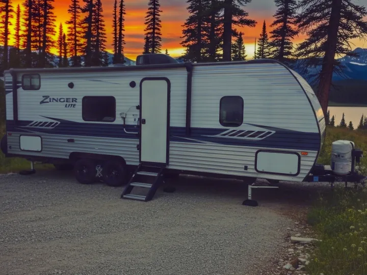 Zinger Lite RV rental parked on gravel, beside a lake with tall pines & mountains under a vibrant sunset. Ideal for camping.. Crossroads RV Zinger Lite 2020