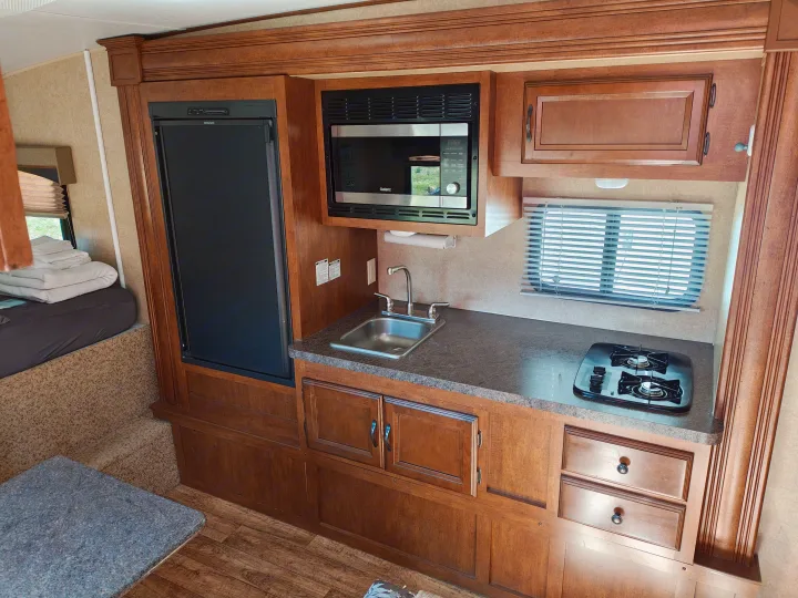 RV rental interior: compact kitchen featuring a Dometic fridge, Galanz microwave, sink, 2-burner stove, wood cabinets & bed area.. Forest River V-Cross Vibe 2012