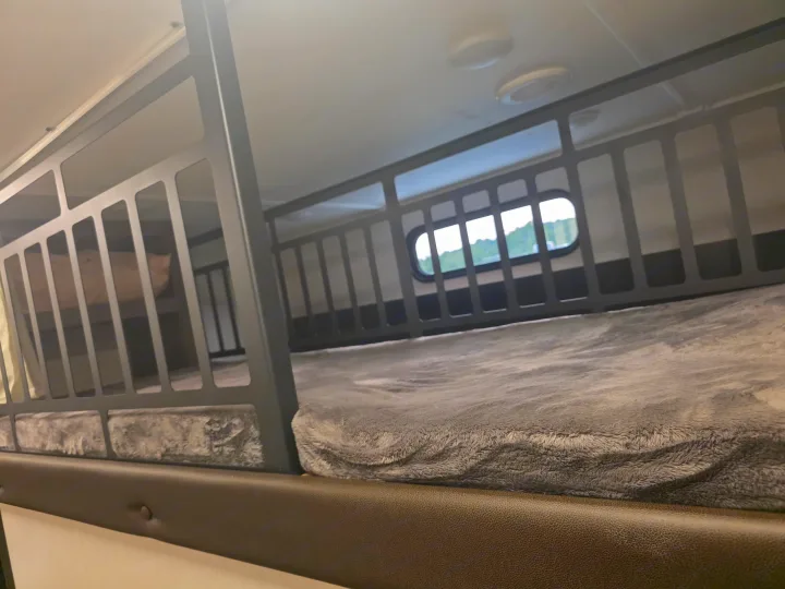 Inside an RV rental, showing compact bunk beds with soft gray blankets. An oval window reveals green scenery.. Forest River Cherokee Alpha Wolf 2026