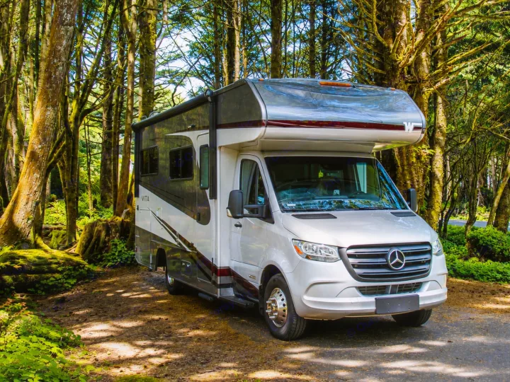 A sleek RV rental, a Mercedes-Benz motorhome, rests amidst tall, mossy forest trees, bathed in dappled sunlight.. Winnebago Vita 2020