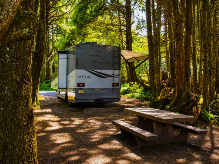 An RV rental motorhome parked under towering trees at a forest campsite with a stone picnic table in dappled sunlight.. Winnebago Vita 2020