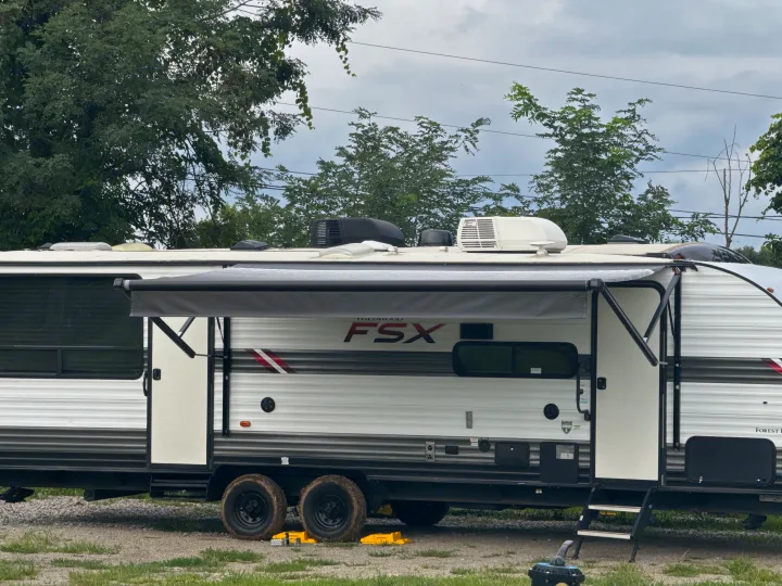 RV rental: A white and gray travel trailer with an extended awning, stairs, and muddy tires, parked on gravel.. Forest River Wildwood FSX Toy Hauler 2021