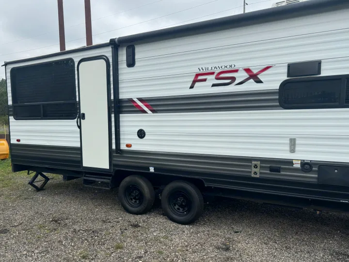 Exterior side of a white Wildwood FSX travel trailer, featuring an awning, windows, and utility hookups. Excellent for rv rental.. Forest River Wildwood FSX Toy Hauler 2021