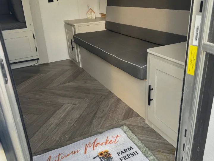 Cozy RV rental interior showcasing herringbone flooring, comfortable seating, cabinets, and a welcoming "Autumn Market" mat.. Forest River Wildwood FSX Toy Hauler 2021