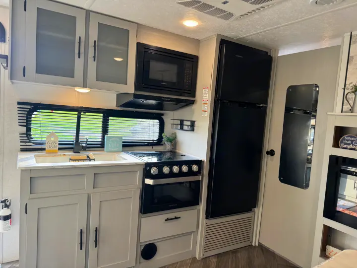 Modern RV rental kitchen featuring gray cabinets, black appliances (oven, microwave, fridge), sink, and window.. Forest River Wildwood FSX Toy Hauler 2021