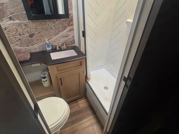 A compact RV rental bathroom with a toilet, vanity, sink, and shower featuring white herringbone tile walls. Grey marble-look backsplash.. Forest River Evo 3005 2024