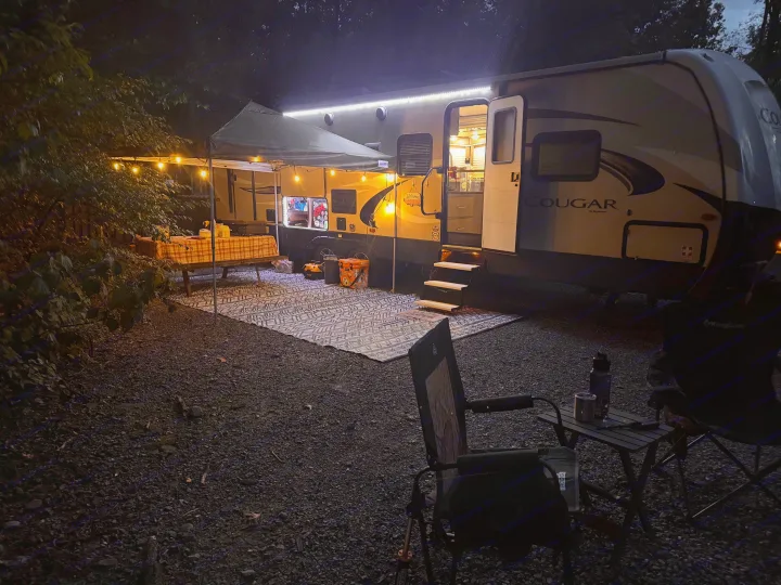 Experience comfort with this rv rental! A lit travel trailer, awning, string lights, and outdoor camp setup at night.. Keystone RV Cougar 2019