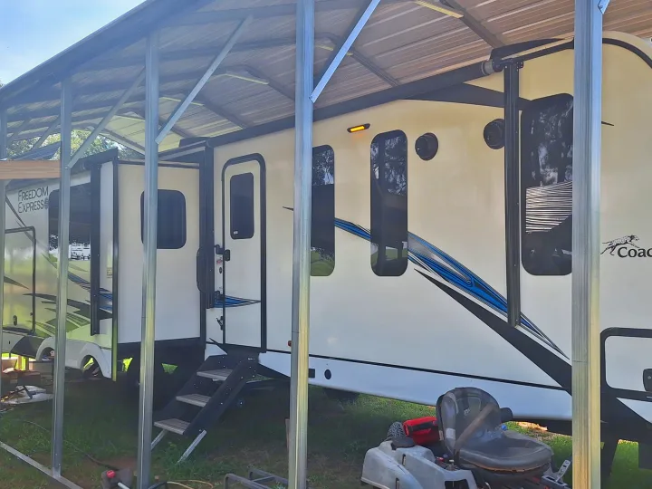 This white Freedom Express rv rental is parked under a protective carport with steps down to the ground. A mower is nearby.. Coachmen Liberty express 2021