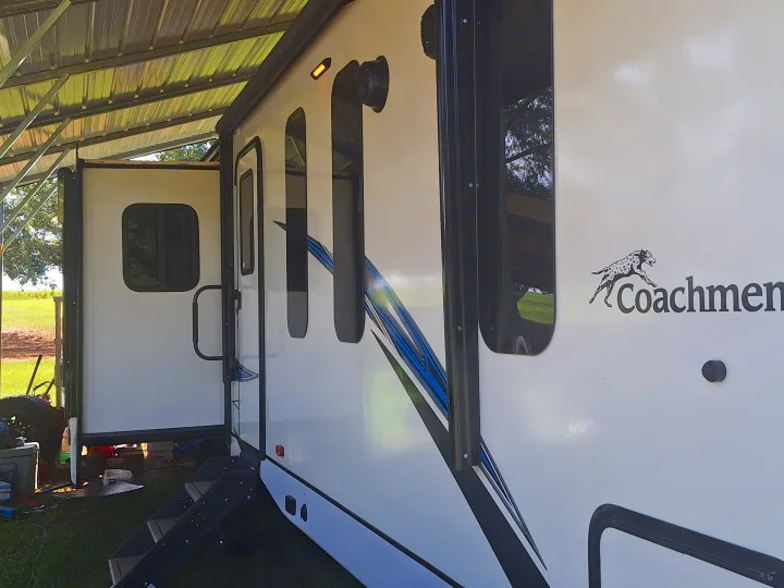 White Coachmen RV rental parked under a metal canopy, showing its side with steps, windows, and graphic stripes.. Coachmen Liberty express 2021
