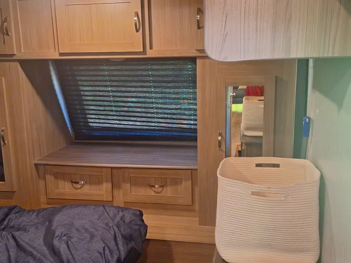 RV rental bedroom with light wood storage cabinets, window blinds, a bed, reflected mirror view, and a white rope basket.. Coachmen Liberty express 2021