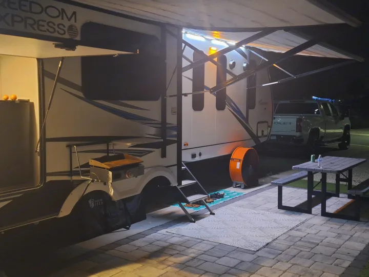 A cozy RV rental campsite at night with an extended, lit awning, outdoor grill, fan, and picnic table ready for relaxation.. Coachmen Liberty express 2021