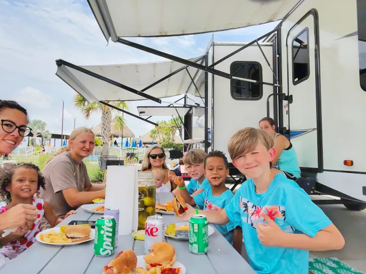 A joyful group enjoys an outdoor meal with burgers, hot dogs, and drinks at their RV rental campsite.. Coachmen Liberty express 2021