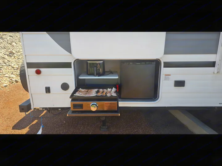 RV rental outdoor kitchen with a griddle cooking bacon, a mini-fridge, and an air fryer, perfect for convenient camping meals.. Forest River Wildwood X-Lite 2025