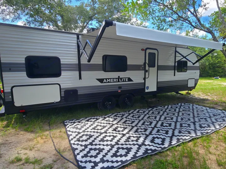 This RV rental, an Ameri Lite travel trailer, is parked at a campsite with its awning extended and a patterned outdoor rug laid out.. Gulf Stream Ameri-Lite 2024