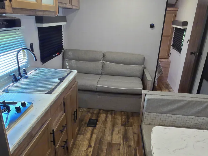 Cozy RV rental interior: kitchen with stove & sink, comfy sofa, and dinette. Modern light wood cabinets, gray walls, and wood-look floor.. Gulf Stream Ameri-Lite 2024