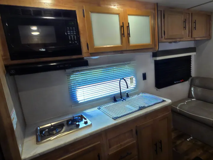 Modern RV rental kitchen featuring a microwave, stovetop, sink, wood cabinets, and window with blinds, plus a seating area.. Gulf Stream Ameri-Lite 2024