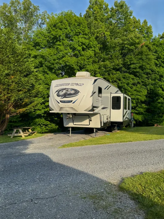A spacious Arctic Wolf Cherokee fifth-wheel RV rental is parked on a sunny gravel campsite with a picnic table and lush trees.. Forest River Cherokee Arctic Wolf 2022