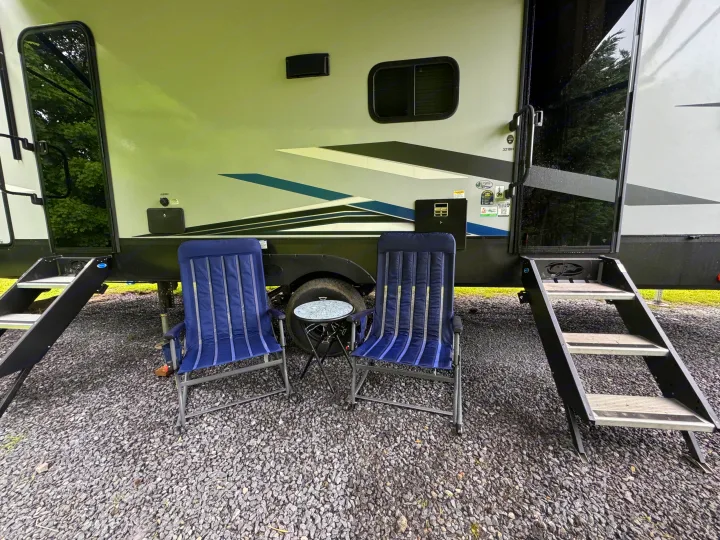 RV rental setup: a parked camper on gravel, flanked by two blue camping chairs, a small table, and entry steps.. Forest River Cherokee Arctic Wolf 2022