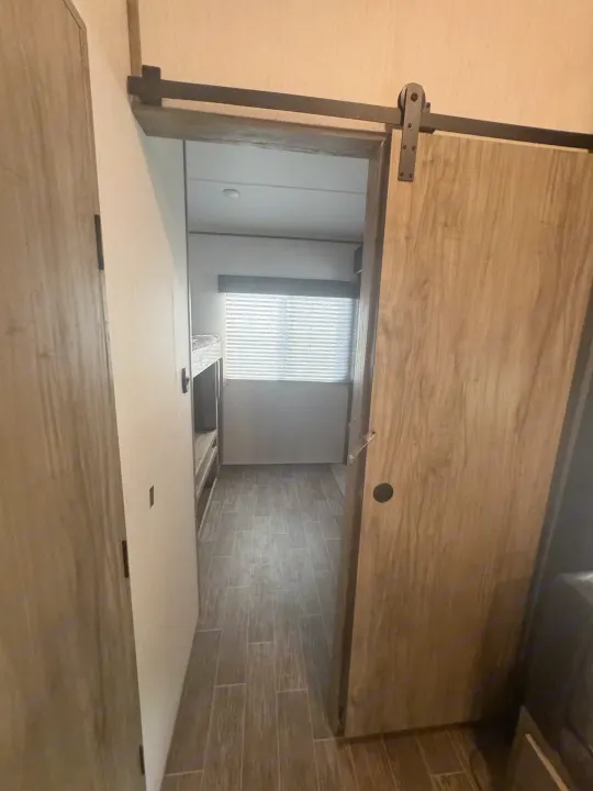 This RV rental interior shows a narrow hallway leading to a bunkhouse, featuring a sliding barn door, wood-look flooring, and a window.. Forest River Cherokee Arctic Wolf 2022