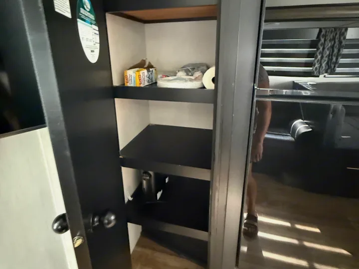 RV rental pantry: dark shelves hold Glad bags, paper plates, and paper towels. Reflection in the fridge shows a person.. Forest River Cherokee Grey Wolf 2022