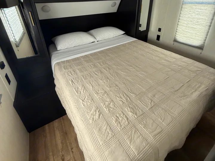 Cozy RV rental bedroom features a bed with white pillows, a beige quilted comforter, a mirror, and a window with blinds.. Forest River Cherokee Grey Wolf 2022
