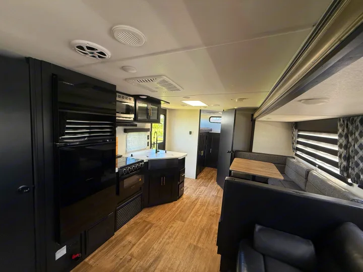 Modern rv rental interior: dark kitchen with fridge, oven, sink; light wood-look floor, dining booth, and comfy sofa.. Forest River Cherokee Grey Wolf 2022