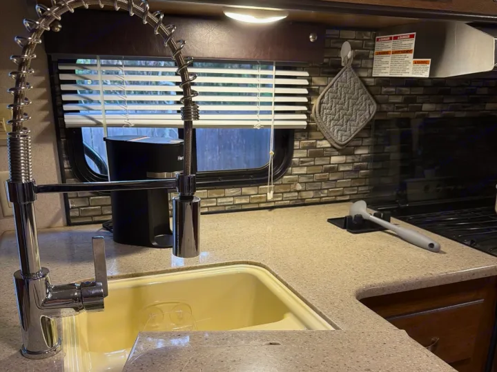 Modern chrome faucet over a light-colored sink in an RV rental kitchen, featuring a tiled backsplash, window, and stove area.. Forest River T27TDSS 2017
