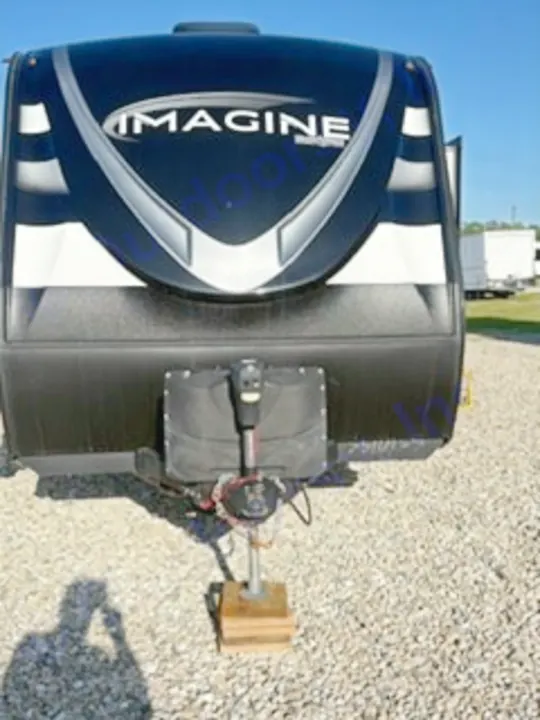 Front view of a black & white 'Imagine' travel trailer, perfect for an RV rental. Visible hitch, propane tanks, and a shadow on gravel.. Grand Design Imagine 2023