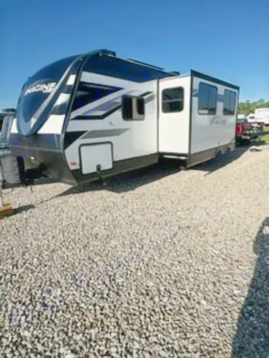 This Imagine travel trailer, perfect for your next RV rental, sits on a gravel lot under a sunny sky, ready for camping adventures.. Grand Design Imagine 2023