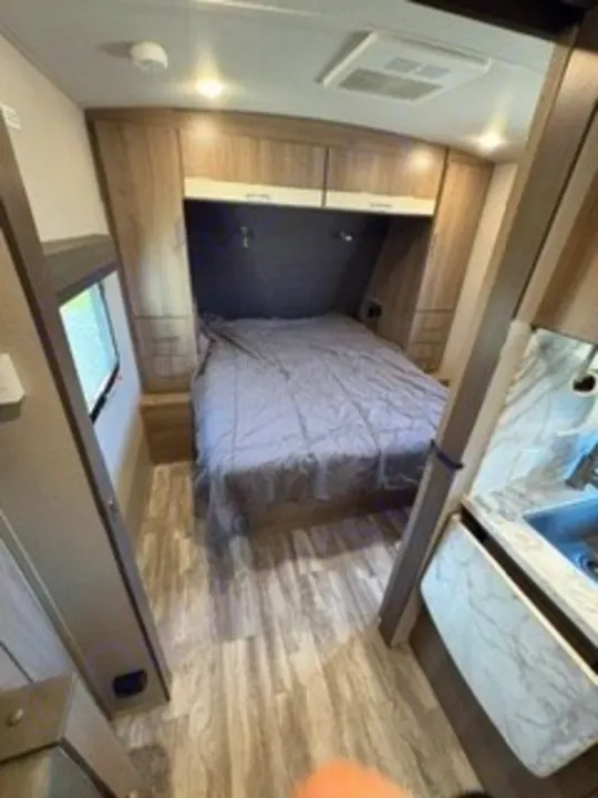 RV rental interior: A modern bedroom with a grey bed, overhead cabinets, wood-look floor, and a partial kitchen view.. Grand Design Imagine 2023