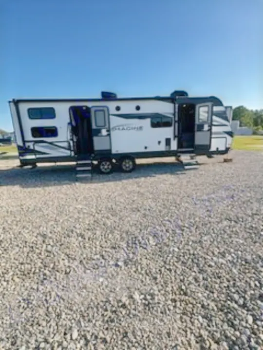 Ready for adventure? This white travel trailer RV rental, with open doors and steps on a gravel lot, awaits your next trip.. Grand Design Imagine 2023