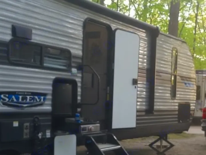 An RV rental travel trailer is set up at a scenic, wooded campsite with outdoor mats. Sunlight brightens the green trees.. Forest River Salem 2022