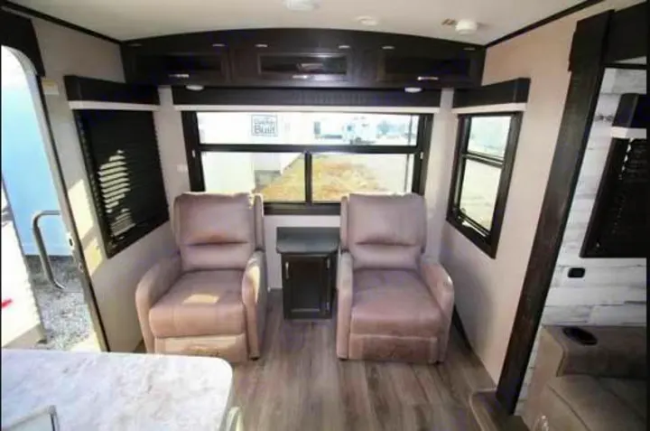 RV rental interior: A bright living area with two plush recliners, end table, large windows, and dark overhead cabinets for comfy trips.. Jayco White Hawk 2021