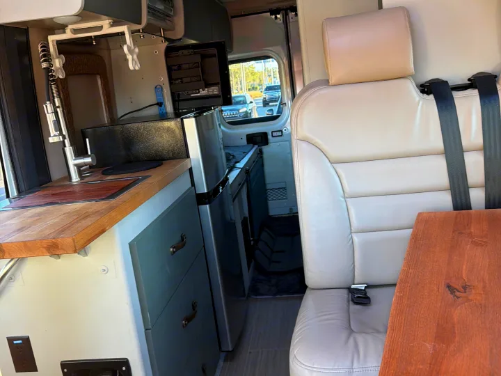 RV rental interior: cozy kitchenette with sink and fridge, plus a beige passenger seat and a wooden table.. Dodge 3500shc 2021
