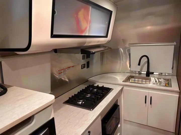 Compact and modern kitchen in an rv rental, featuring a gas stovetop, sink, microwave, and sleek aluminum interior.. Airstream Caravel 2023